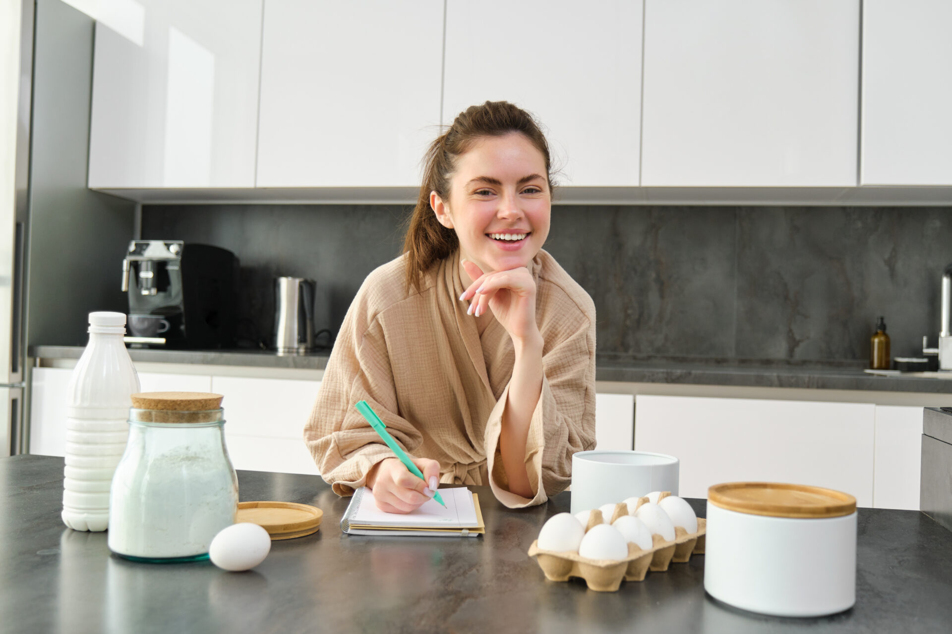 Attractive young cheerful girl baking at the kitchen, making dough, holding recipe book, having ideas.